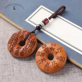 Peach Wood Bagua Coin – two carved coins with Taoist spells and Bagua symbols, brown cord bead accents, blue book backdrop, Daoist shield