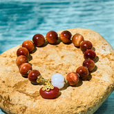 Red Tiger Eye Pumpkin Bracelet – Red tiger eye beads with white pumpkin and red charm on stone, turquoise water background