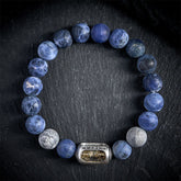 Sodalite Grey Jasper Om Bracelet – top-down view on dark slate, blue beads with metal charm, sodalite healing bracelet