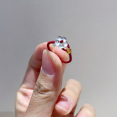 Red String Flower Ring – hand holding red cord ring with white jade flower and gold coin charm, gray background, benming year jewelry