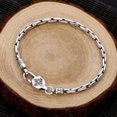 Tibetan Double Dorje Om Mani Padme Hum Bracelet – Silver oval link chain with geometric toggle clasp on wood slice, dark woven mat, top-down flat lay