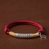 Burgundy cord bracelet with antique silver Tibetan mantra cylinder bead, natural hemp wrapping and lucky fortune coin charm on brown leather background