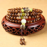 Tibetan Rosewood Bodhi Seed Mala Bracelet with green jade accent beads and carved guru bead, displayed on carved wooden stand, front view, traditional 108-bead meditation necklace wrap bracelet with brown tassel