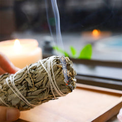 Hand holding lit white sage smudge stick with rising smoke near candle