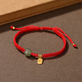 Adjustable red cord bracelet with Hetian jade green bead gold fu blessing fortune charm and sliding knot closure on neutral brown textured background with wooden prop accent