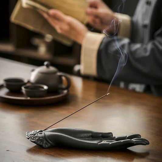 Buddha Hand Incense Burner – matte black ceramic hand holding lit incense stick, smoke rising over blurred tea set, mindful