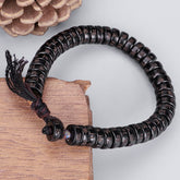 Coconut Shell Om Mani Padme Hum Bracelet – dark carved mantra disc beads on burgundy cord with tassel, pinecone and wood prop