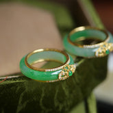 18K Gold Green Chalcedony Strength Ring – Two green jade rings with gold filigree diamond pavé and emerald accents on dark green textured surface, low diagonal