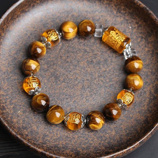 Tiger eye beaded stretch bracelet with carved amber fortune character barrel beads, silver filigree spacer caps and clear crystal dividers in rustic ceramic dish on dark background