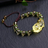 Natural Citrine PiXiu Wealth Bracelet – golden-yellow translucent citrine beads with carved PiXiu wealth charm centerpiece on olive green macrame cord with burgundy adjustable closure, displayed on dark slate surface with natural leaf accents, three-quarter overhead angle
