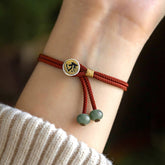 925 Silver Zodiac Natal Buddha Cyan Bodhi Seed Red String Bracelet worn on wrist, braided crimson cord with silver and gold mantra medallion charm, sage green jade terminal beads, lifestyle model shot with knit sweater