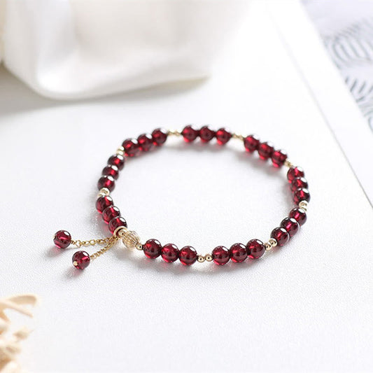 14K gold natural garnet calm bracelet with faceted burgundy garnet beads, gold chain extension with dangling red bead charms, on white fabric with ceramic prop
