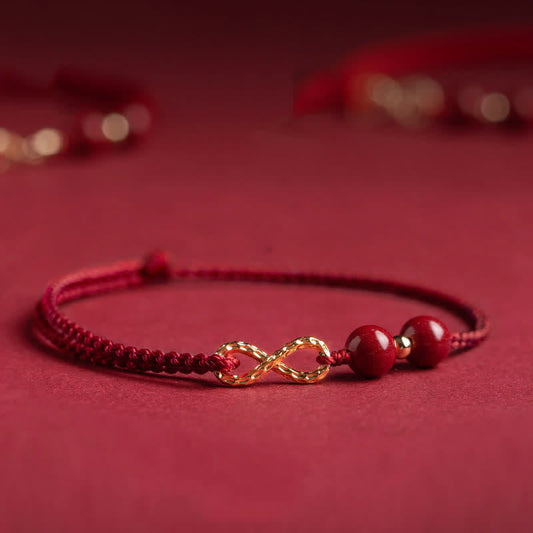Cinnabar Red String 14K Gold Infinity Bracelet flat lay on dusty rose background, braided crimson cord with hammered 14K gold infinity symbol charm, two polished cinnabar beads with gold spacers, adjustable sliding knot