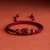 Adjustable burgundy braided cord bracelet with carved cinnabar Om Mani Padme Hum cylindrical bead, gold-toned spacer rings and matching red stone beads on terracotta background