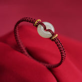 Peace Buckle Jade Abundance Bracelet – Burgundy braided cord with pale celadon jade peace buckle and gold-wrapped knots, crimson fabric background three-quarter