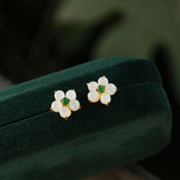 White Jade Flower Blessing Stud Earrings – Five-petal white jade flower earrings with green jade centers in gold settings, dark green velvet jewelry box, elevated side