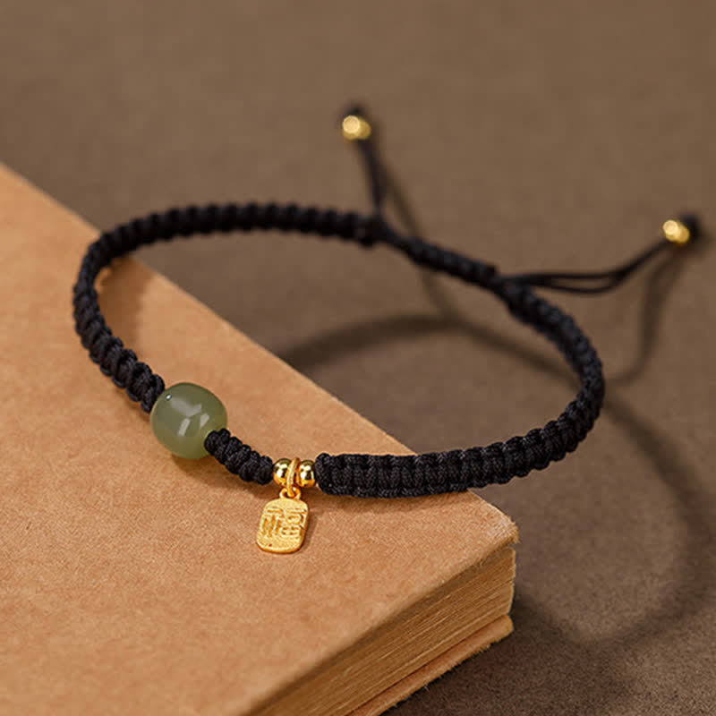 Black cord bracelet with smooth Hetian jade green bead gold-toned rectangular fu fortune charm and gold end caps on aged book surface with warm brown artisanal lifestyle setting
