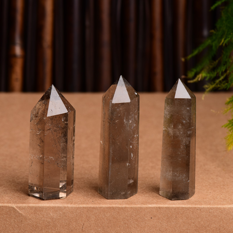Three natural smoky quartz crystal points on brown display stand, bamboo and green plant background, grounding healing towers