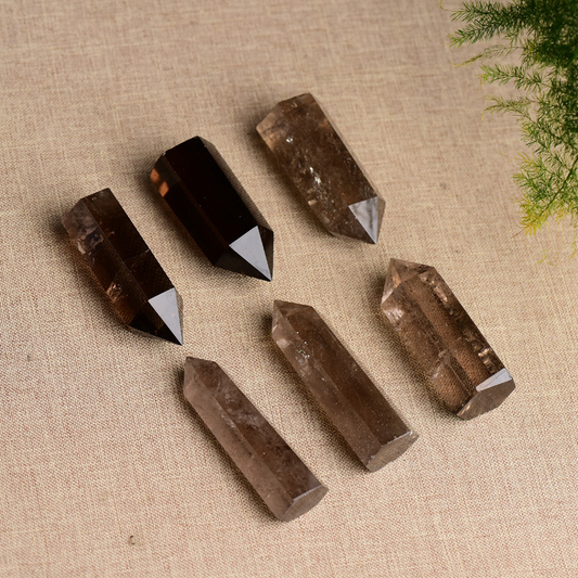 Six natural smoky quartz crystal points arranged on beige fabric, green plant accent, grounding protection stone towers