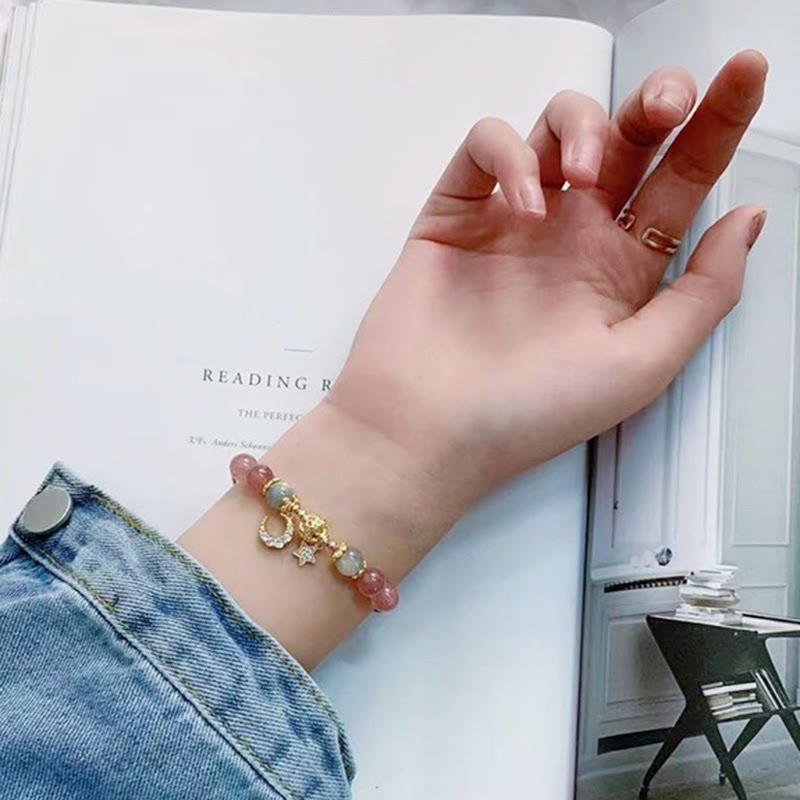 Strawberry Quartz celestial bracelet with pink beads, gray-blue moonstone, gold pavé moon and star charms, cat head charm, styled with open book lifestyle photo.
