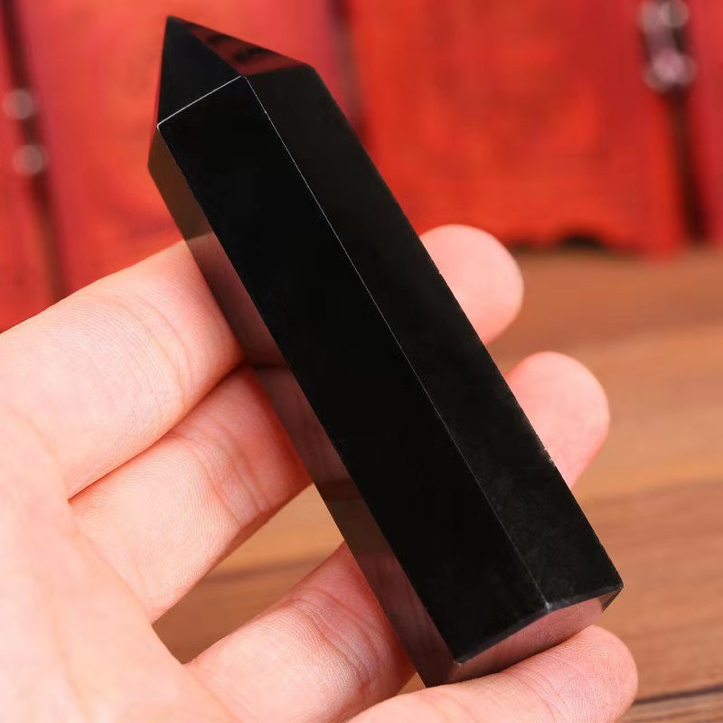Hand holding single black obsidian crystal point tower, close-up side view, red background, grounding protection stone