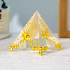 Egyptian Scripture Crystal Pyramid – single clear crystal pyramid with gold hieroglyphic engravings and ornate golden feet, displayed on light linen with soft blue wall background, front view close-up