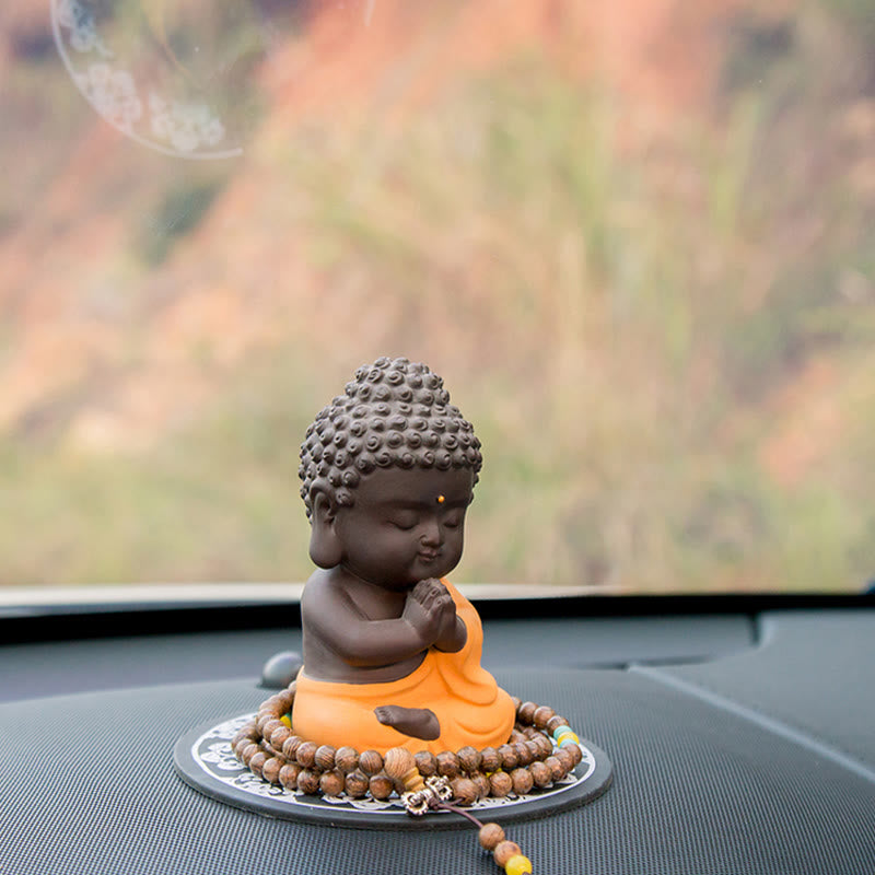 Small Buddha Purple Clay Decoration – orange-robe prayer pose on dashboard with autumn foliage bokeh, seasonal zen driving