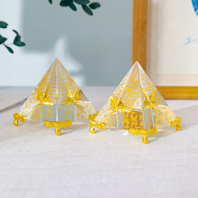 Egyptian Scripture Crystal Pyramid – two clear crystal pyramids with gold hieroglyphic engravings and golden feet side by side on light linen, green leaves and wooden frame in background, lifestyle product shot