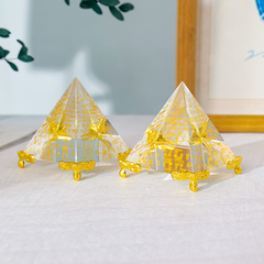 Egyptian Scripture Crystal Pyramid – two clear crystal pyramids with gold hieroglyphic engravings and golden feet side by side on light linen, green leaves and wooden frame in background, lifestyle product shot