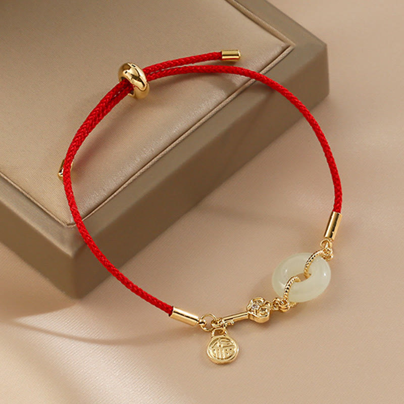 Adjustable red braided cord bracelet with white jade peace buckle pendant, gold fortune Fu character charm and crystal detail on taupe jewelry box