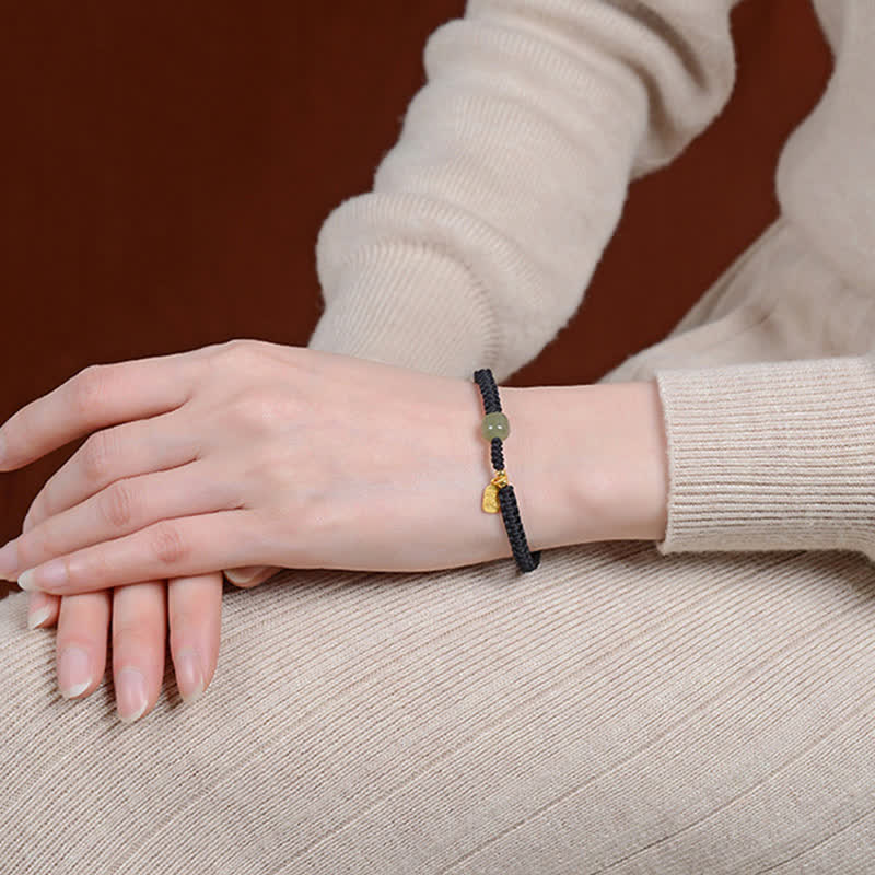 Black cord bracelet with Hetian jade green bead gold fu fortune character charm and gold cord end caps on vintage aged book display with warm artisanal background styling