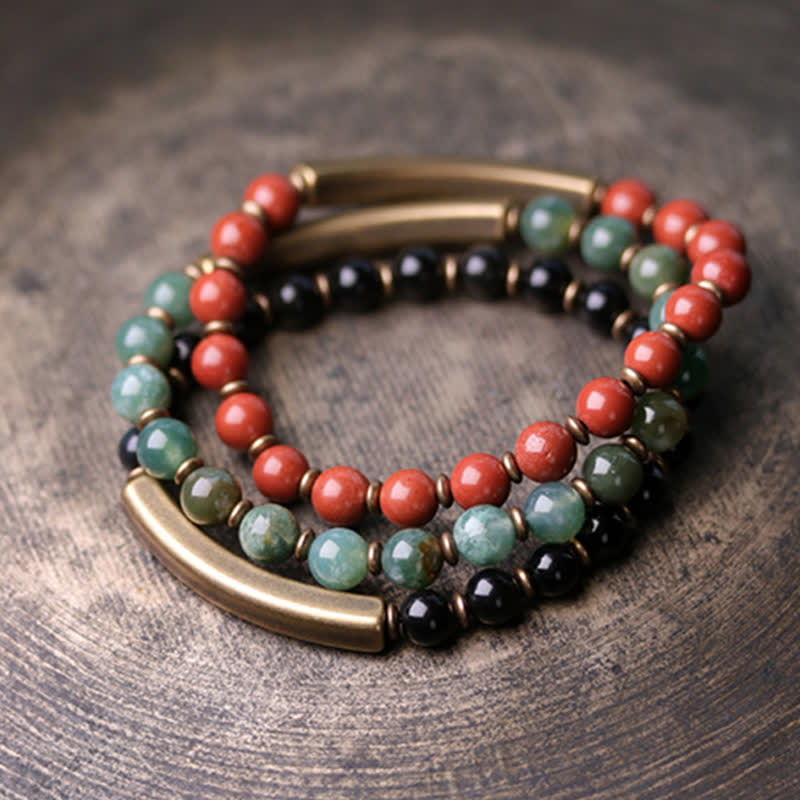 Triple-strand bracelet detail showing red terracotta, green moss agate and black obsidian bead strands with brass bar connectors on rustic wood surface