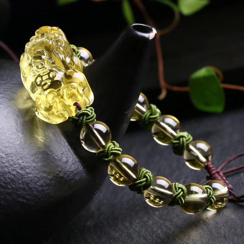 Natural Citrine PiXiu Wealth Bracelet – warm golden citrine beads and carved PiXiu charm on olive green macrame cord with burgundy adjustable tie, styled on dark ceramic vessel with green foliage, low three-quarter angle with soft directional lighting