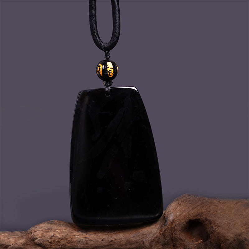 Black Obsidian Dragon Carved Pendant – Trapezoid polished stone with gold mantra bead on gray backdrop, meditative