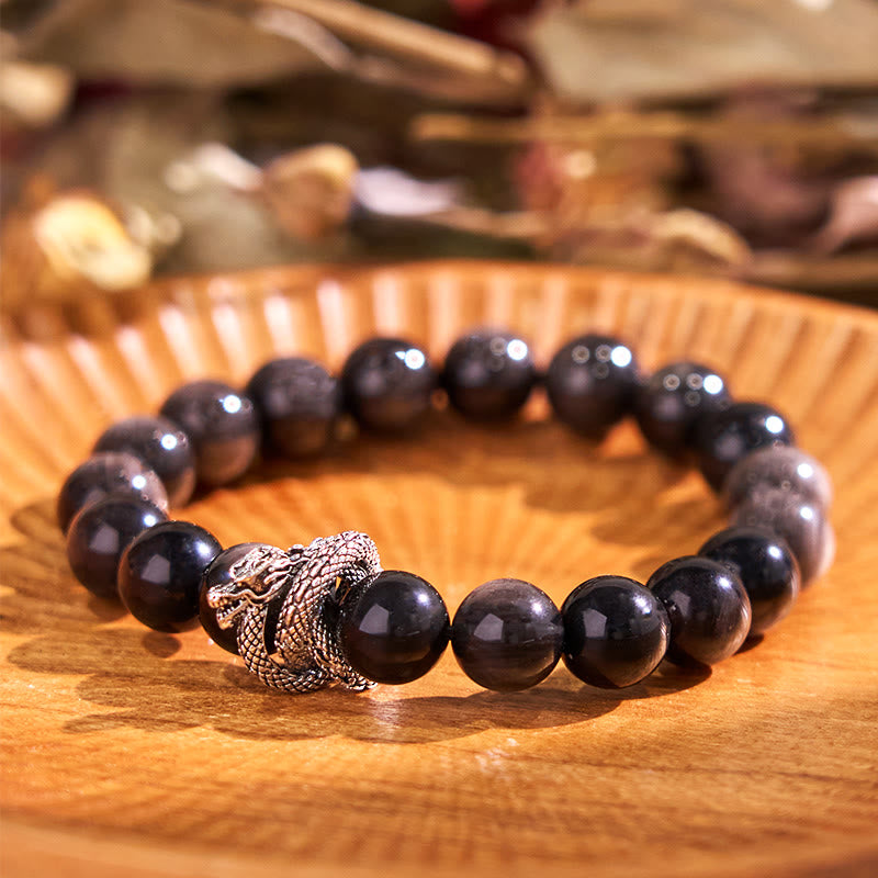 Silver Sheen Obsidian Dragon Bracelet – full bracelet on fluted wooden plate with bokeh background, mystical aura