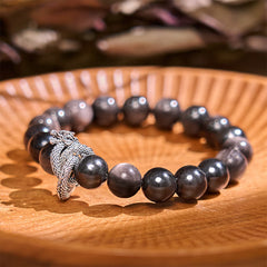 Silver Sheen Obsidian Dragon Bracelet – glossy black beads with silver dragon charm on carved wooden dish, protective energy