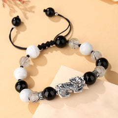 Black Obsidian Pi Xiu Bracelet – Angled flat lay on beige paper, black cord knot detail, sterling silver Pi Xiu centerpiece