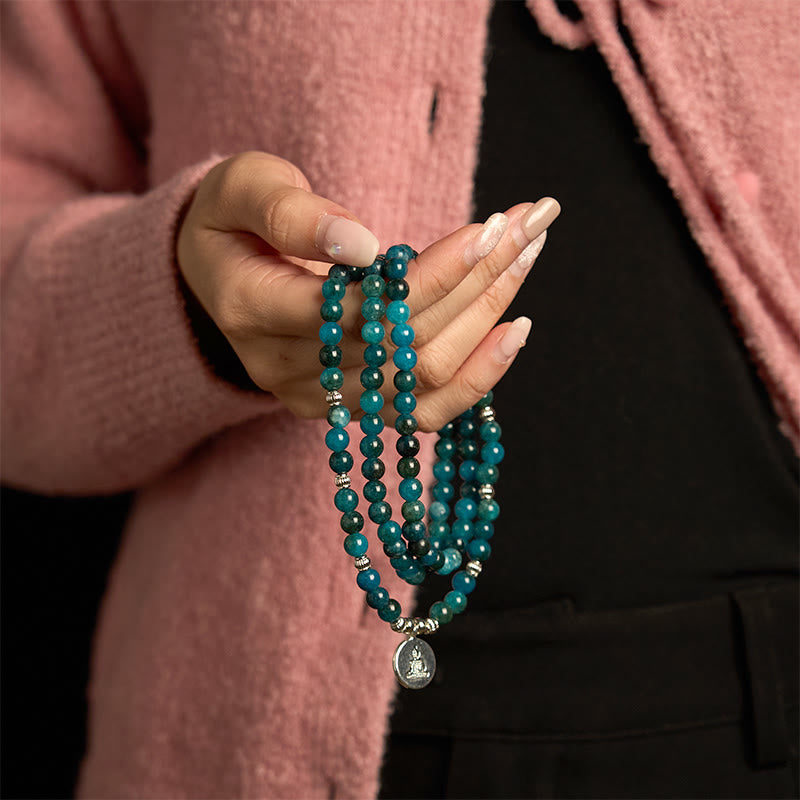 108 Mala Apatite Bracelet - hand holding teal bead strands with Buddha medallion, pink knit cardigan