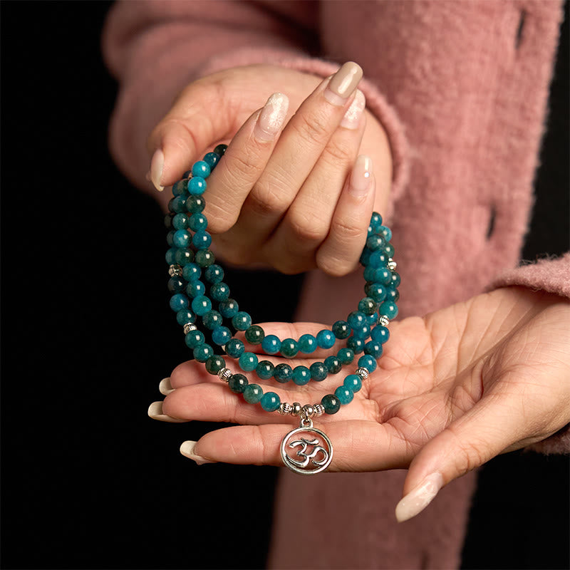108 Mala Apatite Bracelet - two hands displaying multi-strand teal beads with Om charm on dark background
