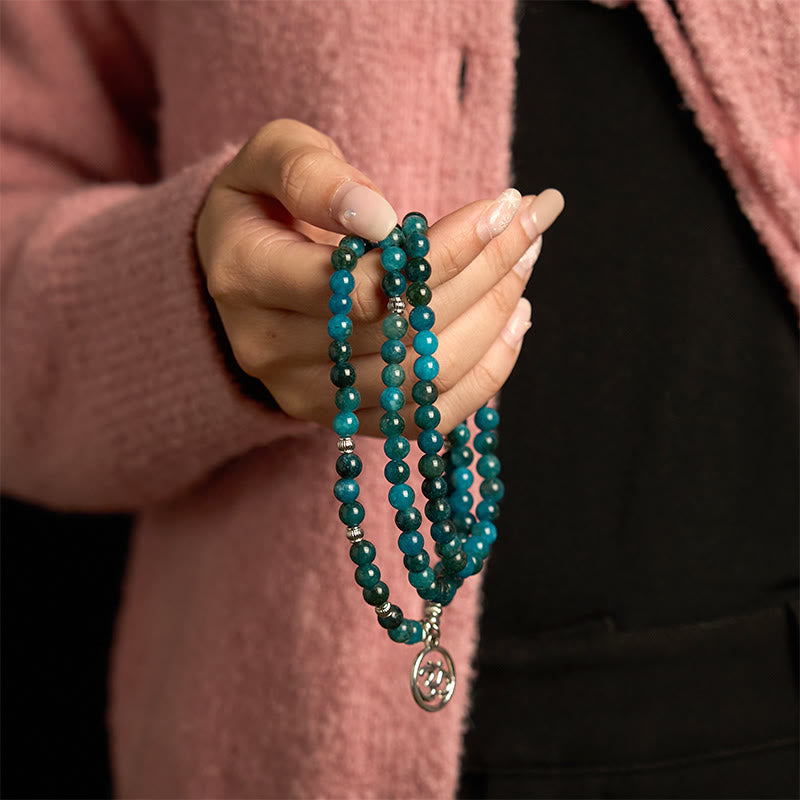 108 Mala Apatite Bracelet - hand holding teal bead strands with Om pendant, pink sweater, black background