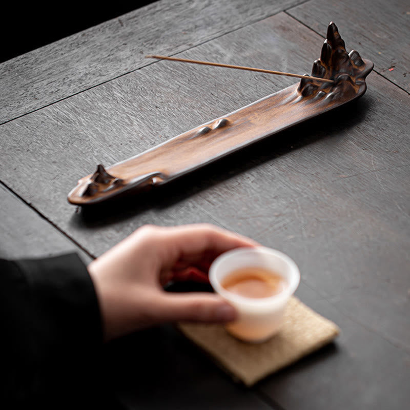 Mountains Boat Ceramic Incense Burner – hand holding tea cup beside brown mountain incense burner on dark wooden table