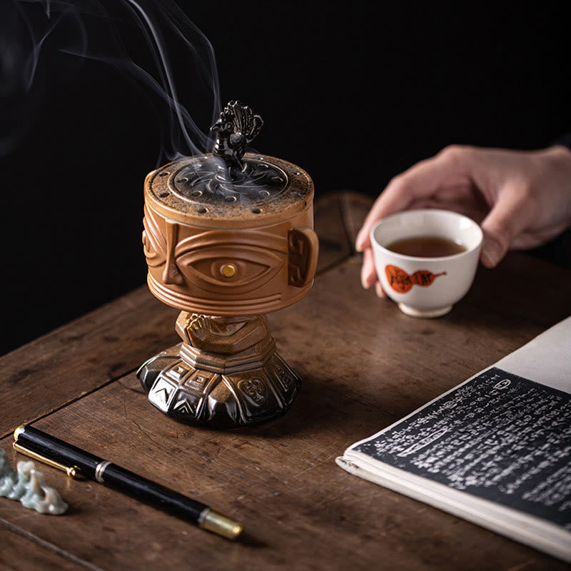 Sanxingdui Ceramic Incense Burner – yellow mask burner beside hand holding teacup over open book, tea ceremony ritual