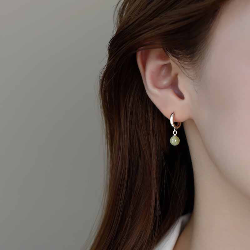 Round Jade Bead Prosperity Earrings – silver huggie hoop with sage green jade drop on model ear, neutral gray background, worn shot