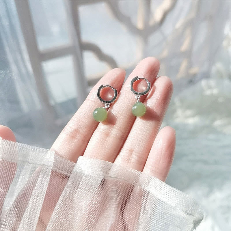Round Jade Bead Prosperity Earrings – silver hoops with pale green jade drops held on fingertips, sheer white fabric and window light backdrop