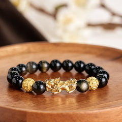 Gold Sheen Obsidian PiXiu Om Mani Padme Hum Wealth Bracelet – Gold PiXiu with obsidian beads on wooden tray, soft floral background