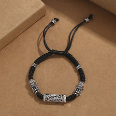 Tibetan PiXiu Bracelet – Black cord bracelet with silver PiXiu and copper Om Mani Padme Hum cylinder on gray surface