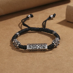 Tibetan PiXiu Bracelet – Black braided cord with silver PiXiu and copper Om Mani Padme Hum cylinder on brown paper