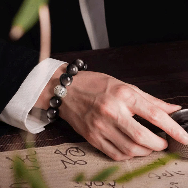 Silver Sheen Obsidian Bracelet – resting hand on dark wood near calligraphy mat and metal dish, meditative grounding