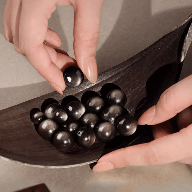 Silver Sheen Obsidian Bracelet – hands holding loose polished black beads in wooden vessel, artisanal grounding