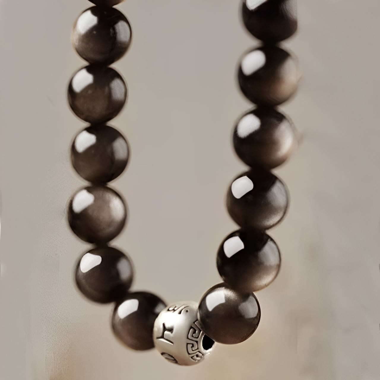 Silver Sheen Obsidian Bracelet – close up draped in U shape on seamless beige background showing mantra bead, grounding